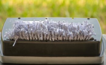 A paper shredder with a jam full of white paper shreds. The paper shredder is near a window that shows grass.