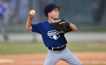 A young baseball athlete pitching in a game. He's mid-throw, wearing gray pants and a navy blue uniform top.