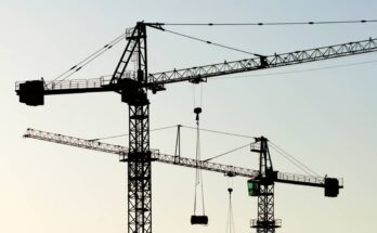 Two large construction cranes hoisted into the air with two pulley systems carrying material. The cranes are black.