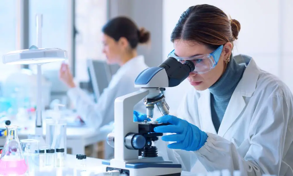 A woman in a lab coat, gloves, and goggles looks at a microscope. Another woman works behind her.