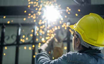 A man wearing a yellow hard hat and denim jacket while grinding steel. Sparks are flying around in front of his face.