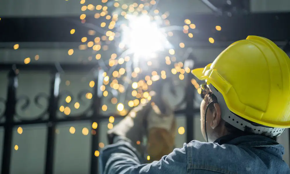 A man wearing a yellow hard hat and denim jacket while grinding steel. Sparks are flying around in front of his face.