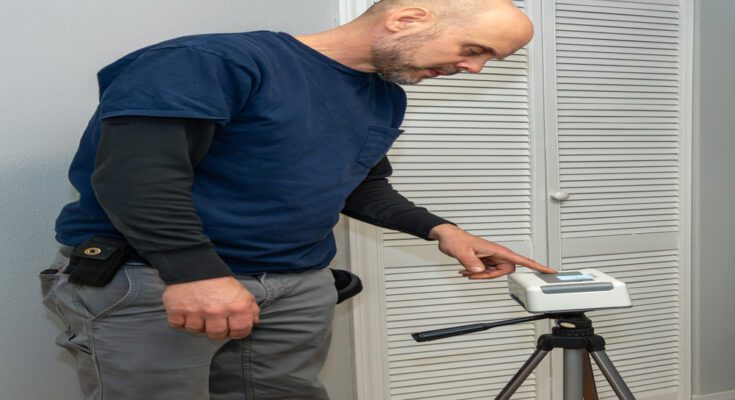 A man wearing a blue shirt and cargo pants sets up a radon test in a room. He is standing near a closet.