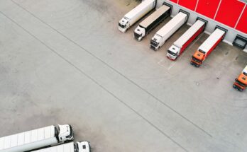 An aerial view shows nine red and white semi-trucks parked at a red warehouse terminal loading area.