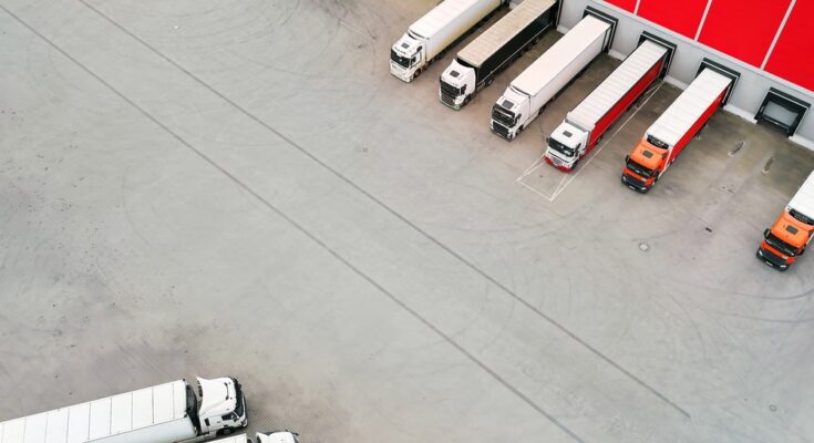 An aerial view shows nine red and white semi-trucks parked at a red warehouse terminal loading area.