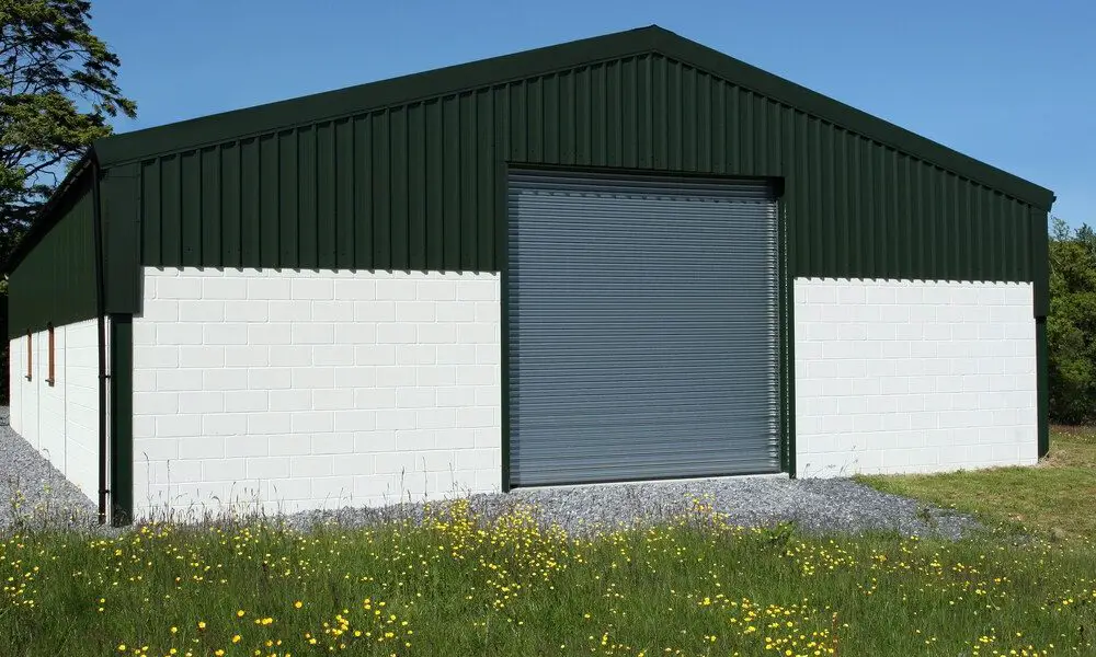 The exterior of a green and white metal barn with a peaked roof and a large gray door. The barn is surrounded by trees.