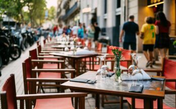 Wooden tables and chairs with red fabric line the sidewalk outside a cafe. There are wine glasses and flowers on the tables.