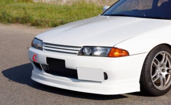 The front bumper, grille, and headlights of a white Nissan Skyline on pavement with grass in the background.