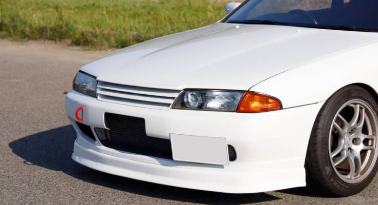 The front bumper, grille, and headlights of a white Nissan Skyline on pavement with grass in the background.