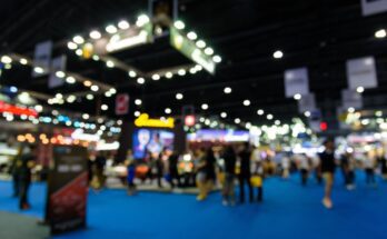 People are walking around a trade show, looking at various booths. The carpet is blue, and the photo is blurry.