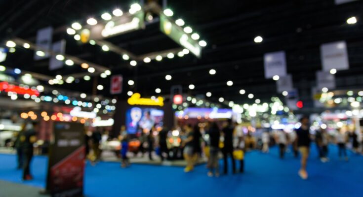 People are walking around a trade show, looking at various booths. The carpet is blue, and the photo is blurry.