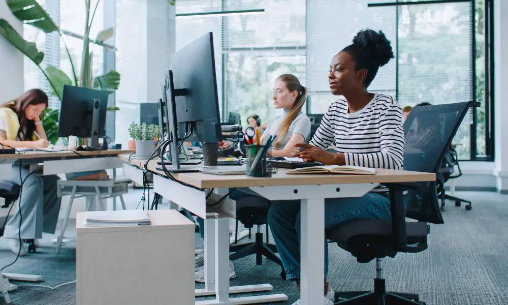 Three women sitting at desks with large monitors and keyboards. The office has tall windows with open blinds.