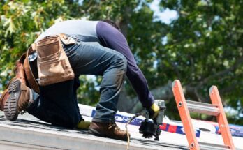 Worker wearing a tool belt using a nail gun on the roof of a home next to an orange ladder with trees in background.