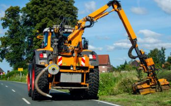 A large, yellow tractor drives down a road while using a boom mower to mow the overgrown vegetation.