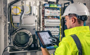An electrical worker wearing a reflective uniform and a white hard hat is holding a tablet in front of a switchboard.