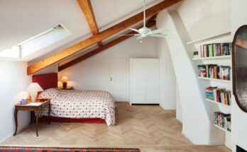 An attic bedroom with a ceiling fan and built-in bookshelf. The room also has exposed wooden beams and a skylight.