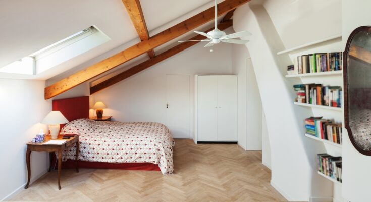 An attic bedroom with a ceiling fan and built-in bookshelf. The room also has exposed wooden beams and a skylight.