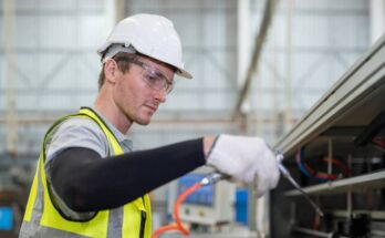 A male engineer with safety goggles cleans a packaging machine using an air duster in a factory warehouse.