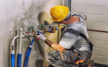A plumber wearing a yellow protective hard hat while using a wrench to work on an industrial plumbing system.