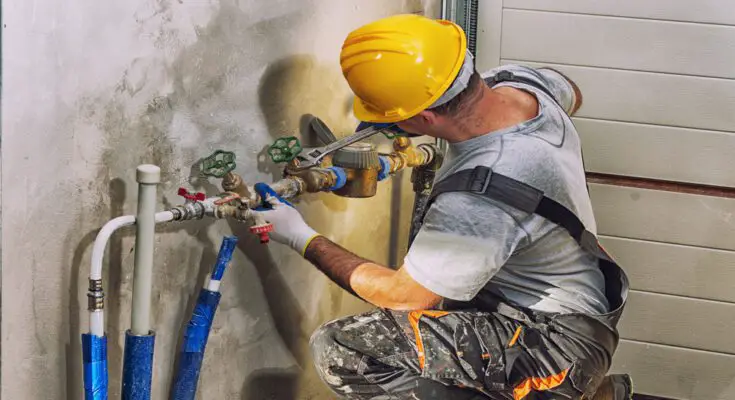 A plumber wearing a yellow protective hard hat while using a wrench to work on an industrial plumbing system.