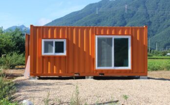 An orange shipping container building with a siding glass door and a window sits in a gravel lot near mountains.