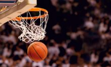 A basketball exiting a swooshing net after scoring a point. In the background is a blurry stand full of spectators.