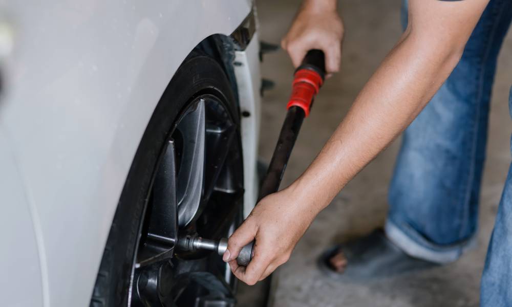 A close-up of a person in jeans and outdoor sandals carefully tightening the lug nuts on a car's wheel.