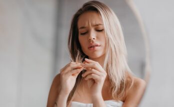 A woman has a white towel wrapped around her body. She is holding a bunch of hair in her hands and looking at it quizzically.