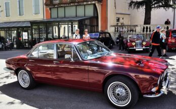 A dark red vintage Jaguar is parked outdoors at a classic car show. The four-door sedan has silver accents and wire wheels.