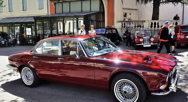 A dark red vintage Jaguar is parked outdoors at a classic car show. The four-door sedan has silver accents and wire wheels.