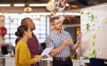 There is a man talking to two people in front of a whiteboard with green sticky notes on it and behind them is a blurred office background.