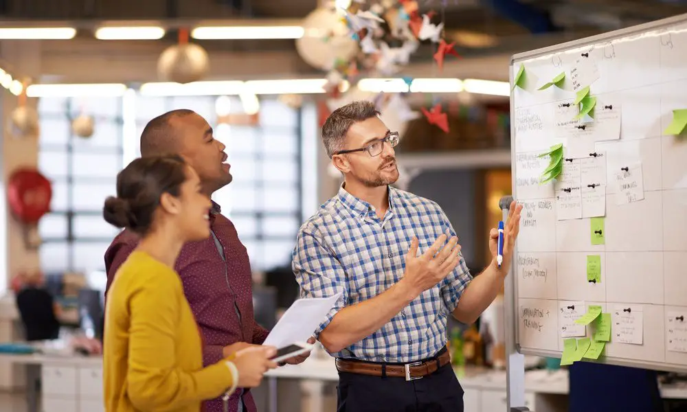 There is a man talking to two people in front of a whiteboard with green sticky notes on it and behind them is a blurred office background.