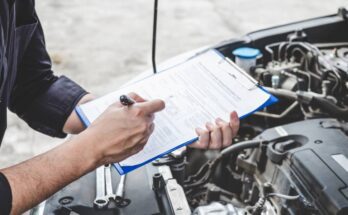 A person looking underneath the hood of a car. They hold a clipboard and a pen to make notes on a document.