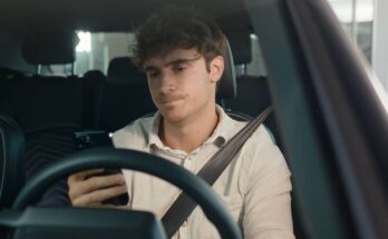 A frustrated looking young man sits in the driver's seat of a vehicle with one hand holding his phone.