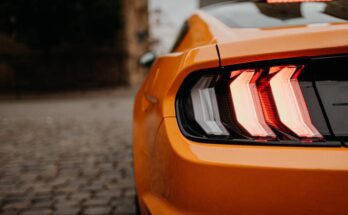 The left rear brake light of an orange Ford Mustang is visible from behind. The light is illuminated and red.