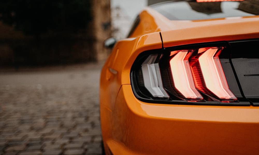 The left rear brake light of an orange Ford Mustang is visible from behind. The light is illuminated and red.