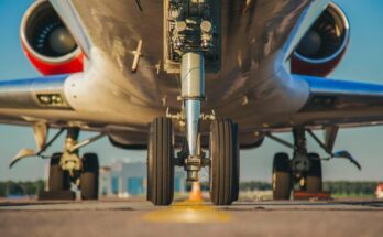 Large chucks hold a plane by its landing gear in place at an airport runway. Buildings are visible in the distance.