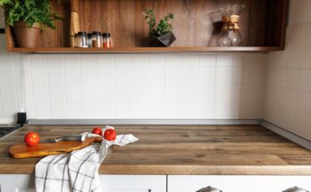 Three red tomatoes, a silver knife, a white cloth, and a wooden cutting board sit on top of a wooden kitchen countertop.
