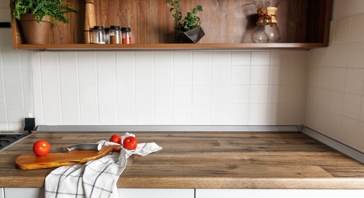 Three red tomatoes, a silver knife, a white cloth, and a wooden cutting board sit on top of a wooden kitchen countertop.