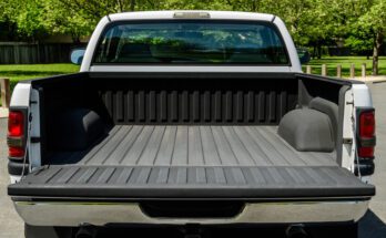 The back of a white truck with the truck bed open and empty, ready for equipment, parked outside of a lush park.