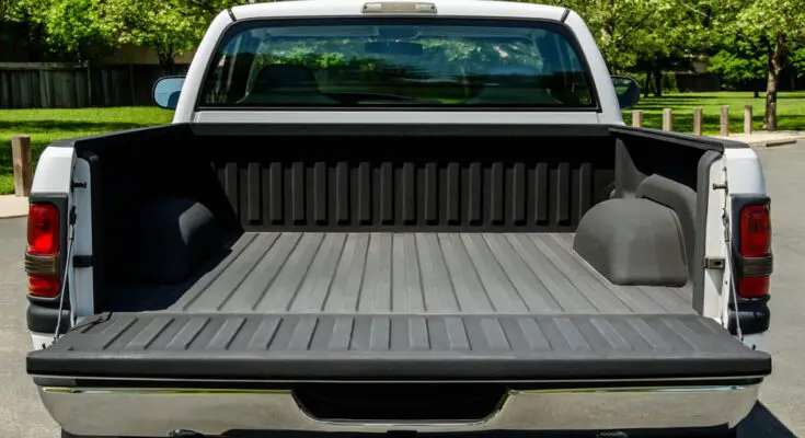 The back of a white truck with the truck bed open and empty, ready for equipment, parked outside of a lush park.