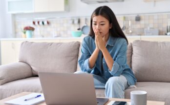 A woman with a concerned facial expression sits on the couch looking at a laptop on the coffee table.