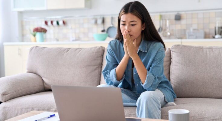 A woman with a concerned facial expression sits on the couch looking at a laptop on the coffee table.