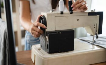 A close-up of a woman using a sewing machine. She is turning a dial at the back of the machine to adjust the settings.