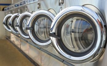 A row of five washing machines, all with their front doors open at the same angle. Each machine is shiny and clean.
