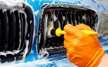 A person with an orange glove using a boar's hair brush to clean and detail the sudsy grill of a blue vehicle.