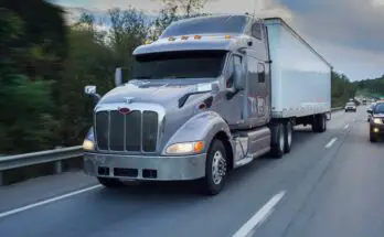 A gray and white semi-truck driving along a tree-lined highway. The truck is surrounded by other vehicles.