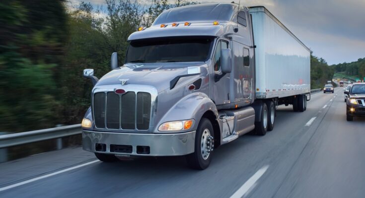 A gray and white semi-truck driving along a tree-lined highway. The truck is surrounded by other vehicles.
