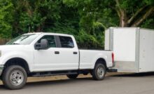 A white pickup truck pulls a white enclosed trailer. The truck and trailer are parked next to large trees.