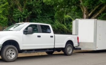 A white pickup truck pulls a white enclosed trailer. The truck and trailer are parked next to large trees.